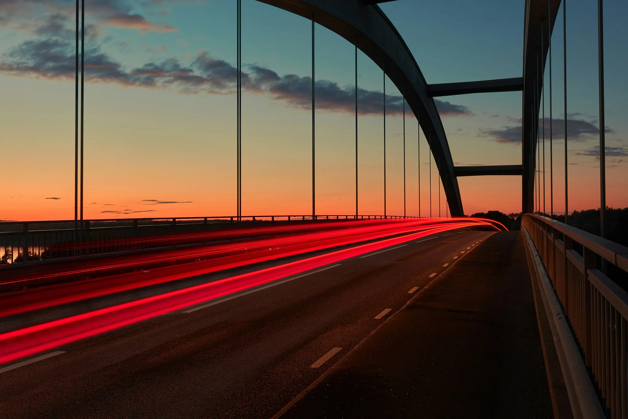 Hängebrücke bei Sonnenuntergang mit roten Fahrzeuglichtspuren und orange-blauem Farbverlauf am Himmel